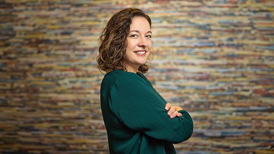 Aron Gelineau stands with her arms crossed and smiles at the camera. 她有一头棕色的卷发，穿着一件蓝绿色的毛衣.