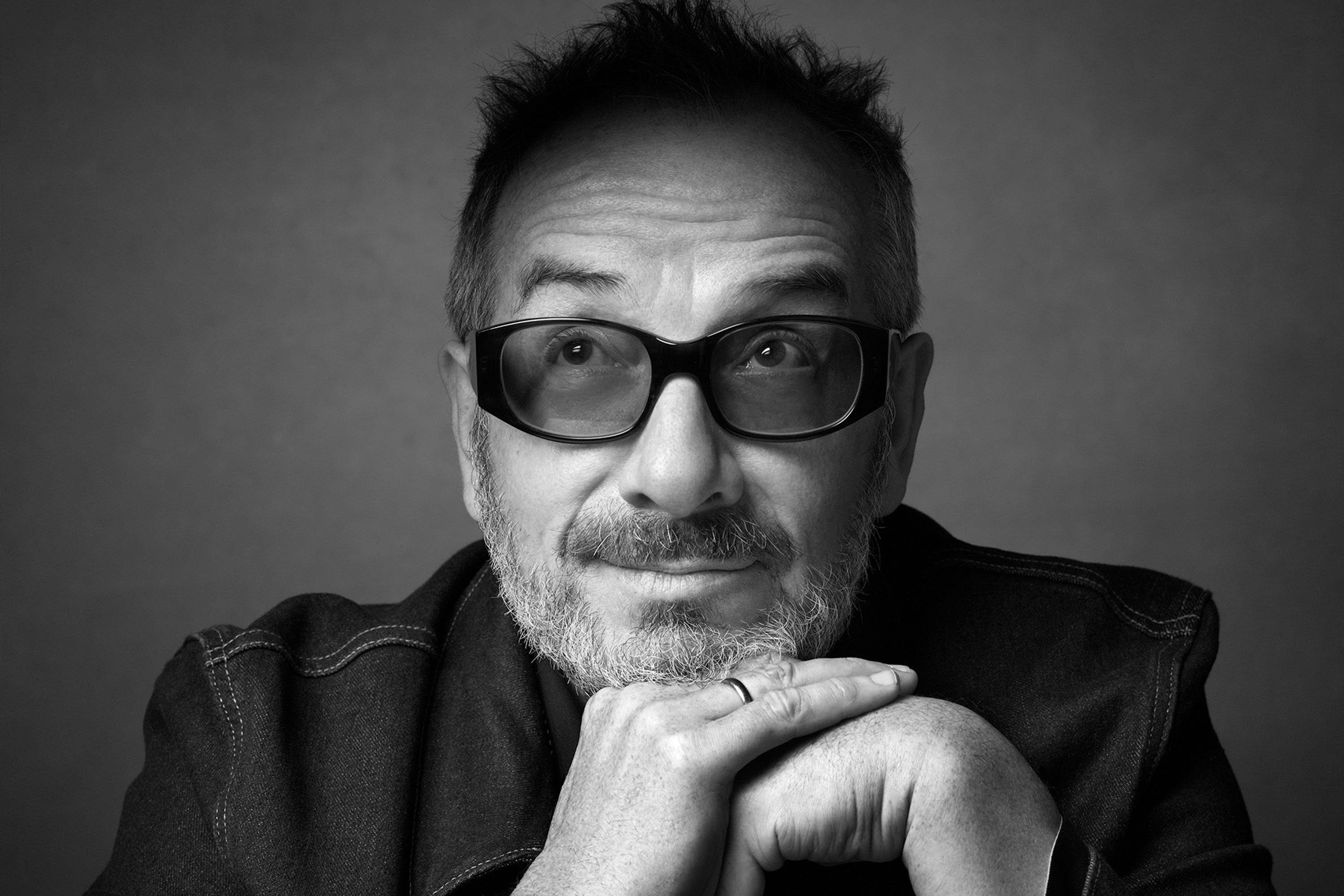 Closeup black and white photo of Elvis Costello with tinted glasses and looking up and to the right with his hands folded under his chin.